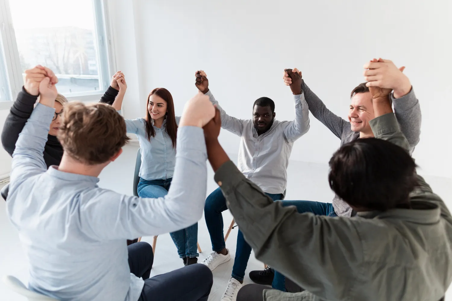 A diverse group of people sitting in a circle, holding hands and smiling, symbolizing unity, teamwork, and support in a collaborative or therapeutic setting.