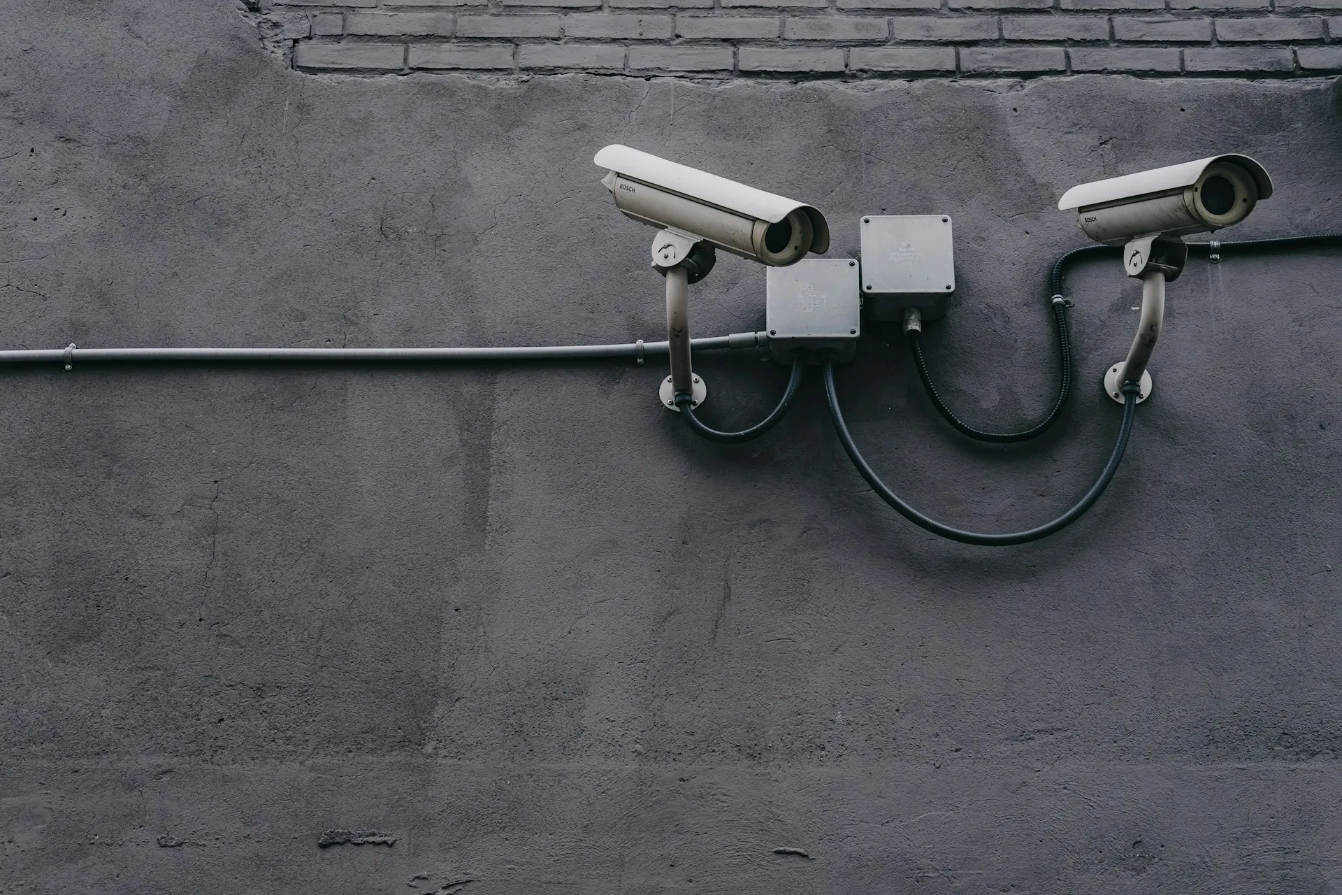 Two security cameras mounted on a gray concrete wall, monitoring the area with visible cables.