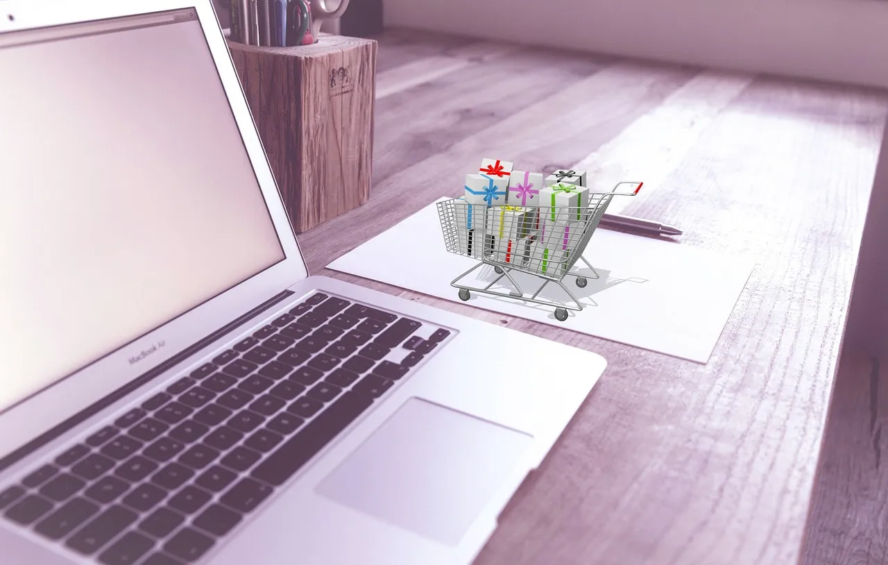 MacBook on wooden desk with miniature shopping cart and gifts.