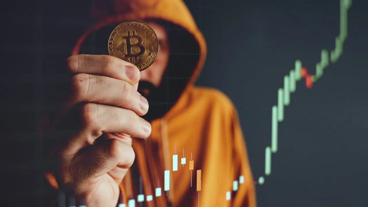 Hooded person holding a Bitcoin coin in front of a rising market chart.