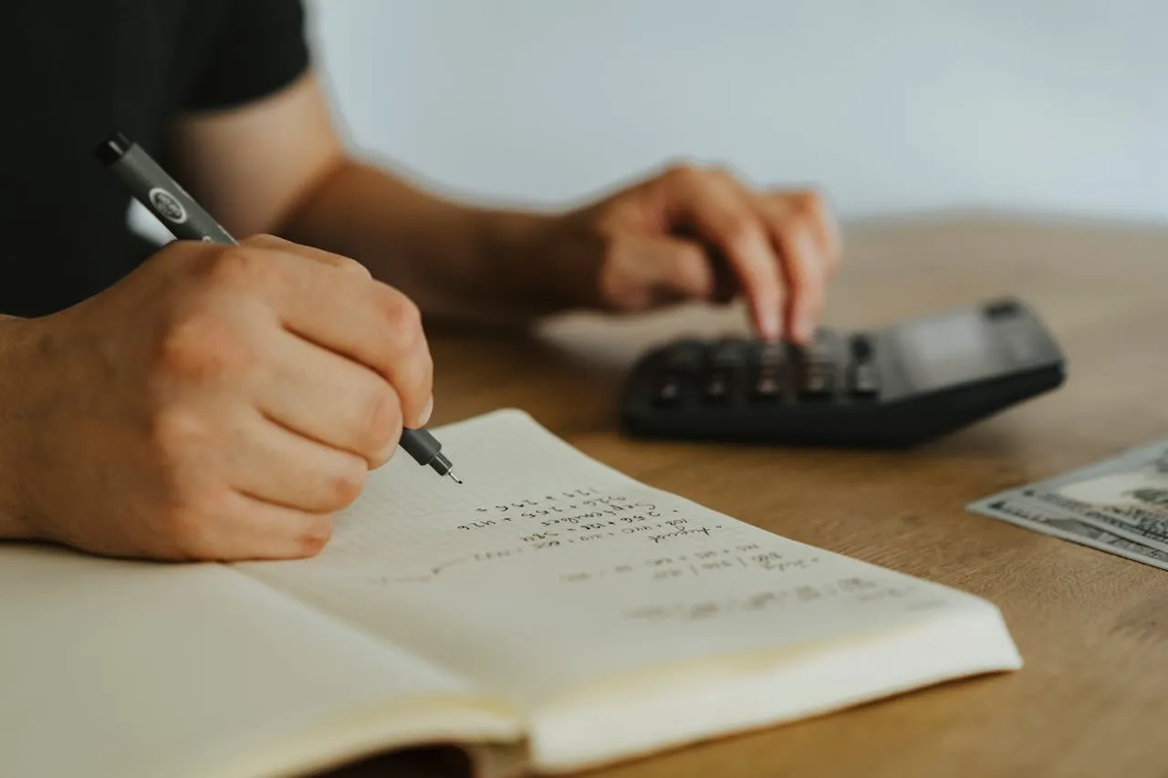 Hands writing financial calculations in notebook with calculator on desk.