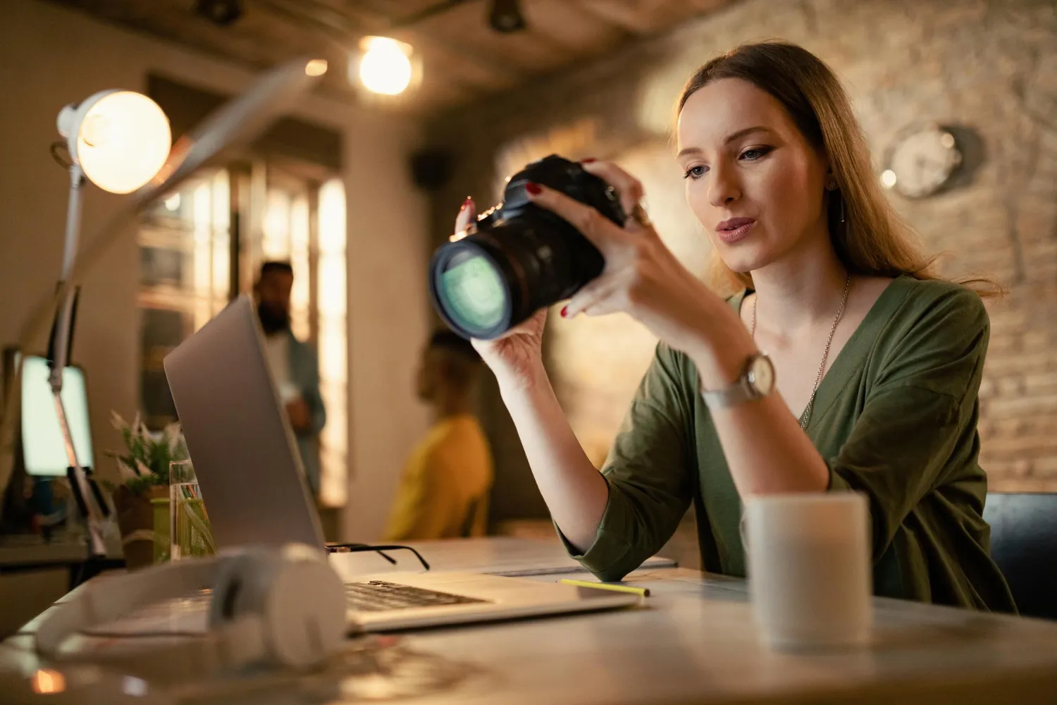 Woman reviewing camera settings at a creative workspace with a laptop, headphones, and coffee cup on the desk.