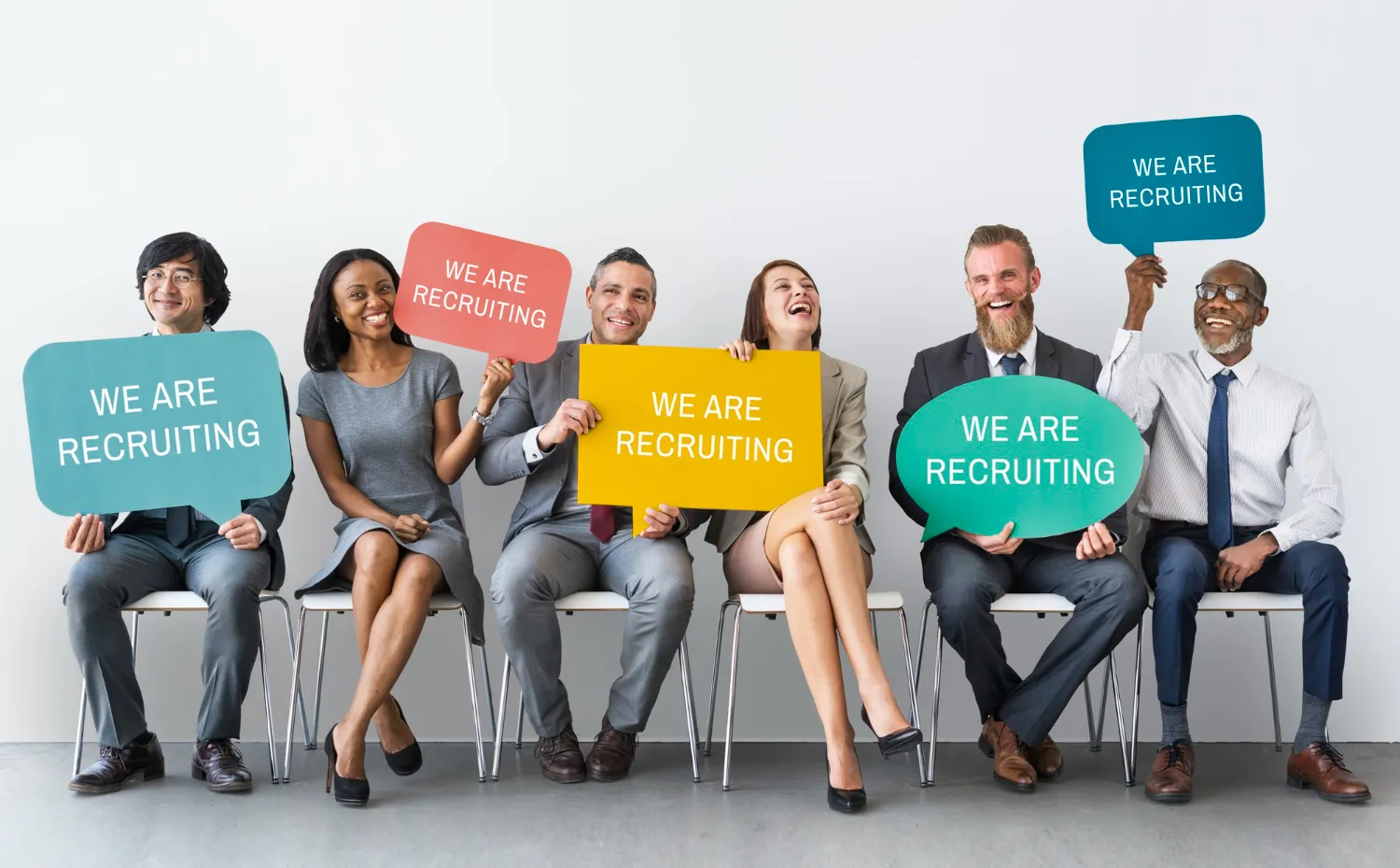 Diverse group of professionals sitting and holding 'We Are Recruiting' signs, promoting hiring and recruitment opportunities.