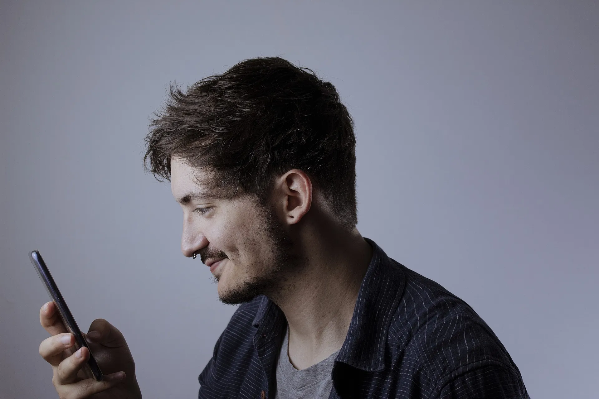 Side profile of a young man smiling while looking at his smartphone screen in a neutral background.