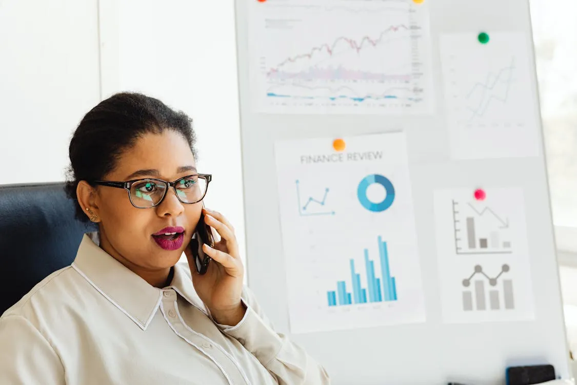 Businesswoman at her desk reviewing financial charts and graphs while talking on the phone; financial data displayed on the board in the background.