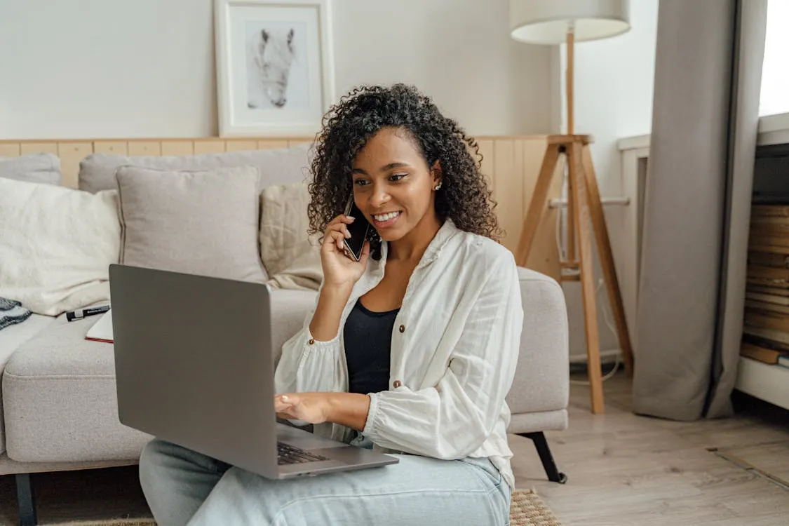 Woman sitting on the floor and working remotely on her laptop while talking on the phone in a cozy living room setting.