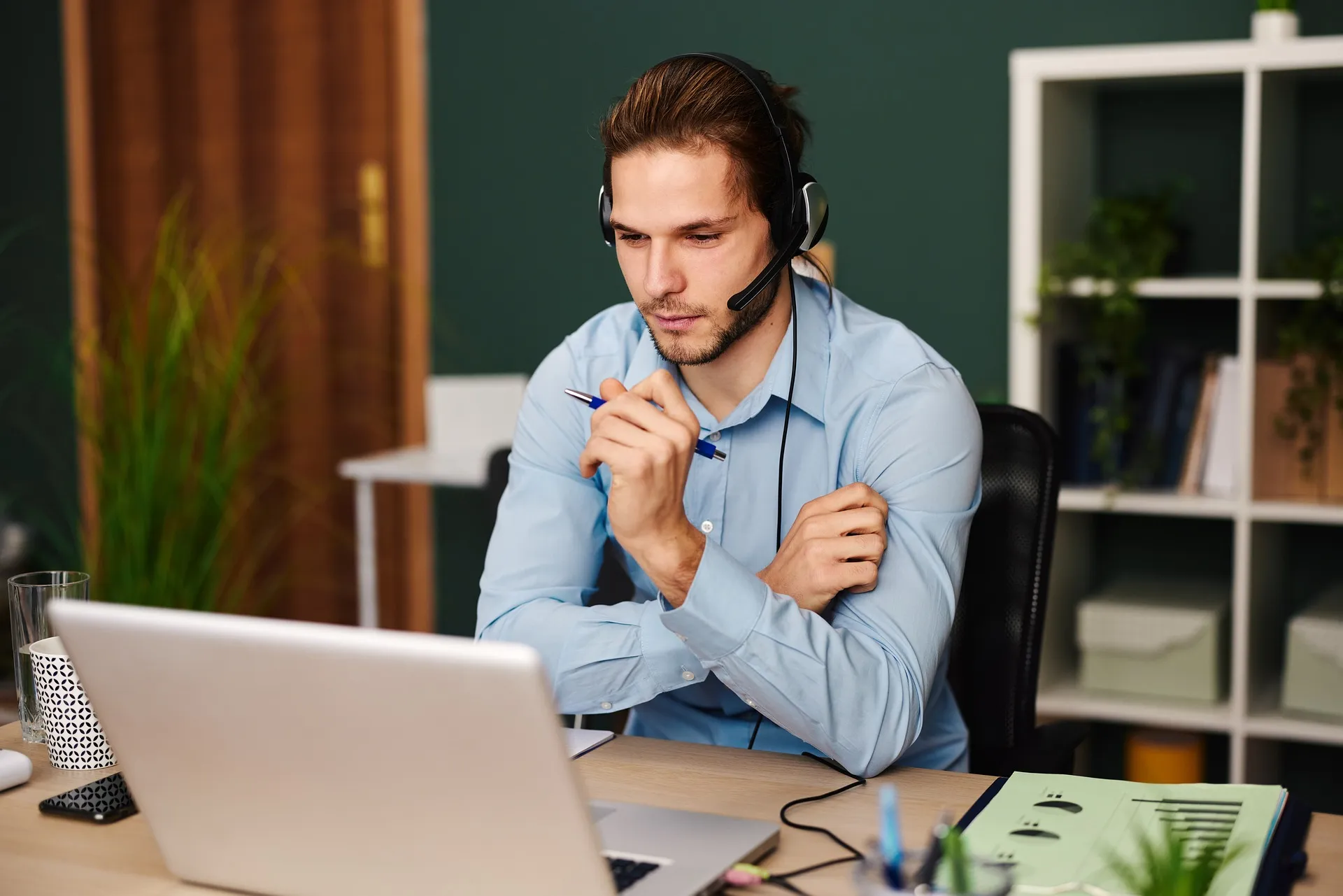 Focused man wearing a headset and looking at a laptop screen during a video call in an office setting.