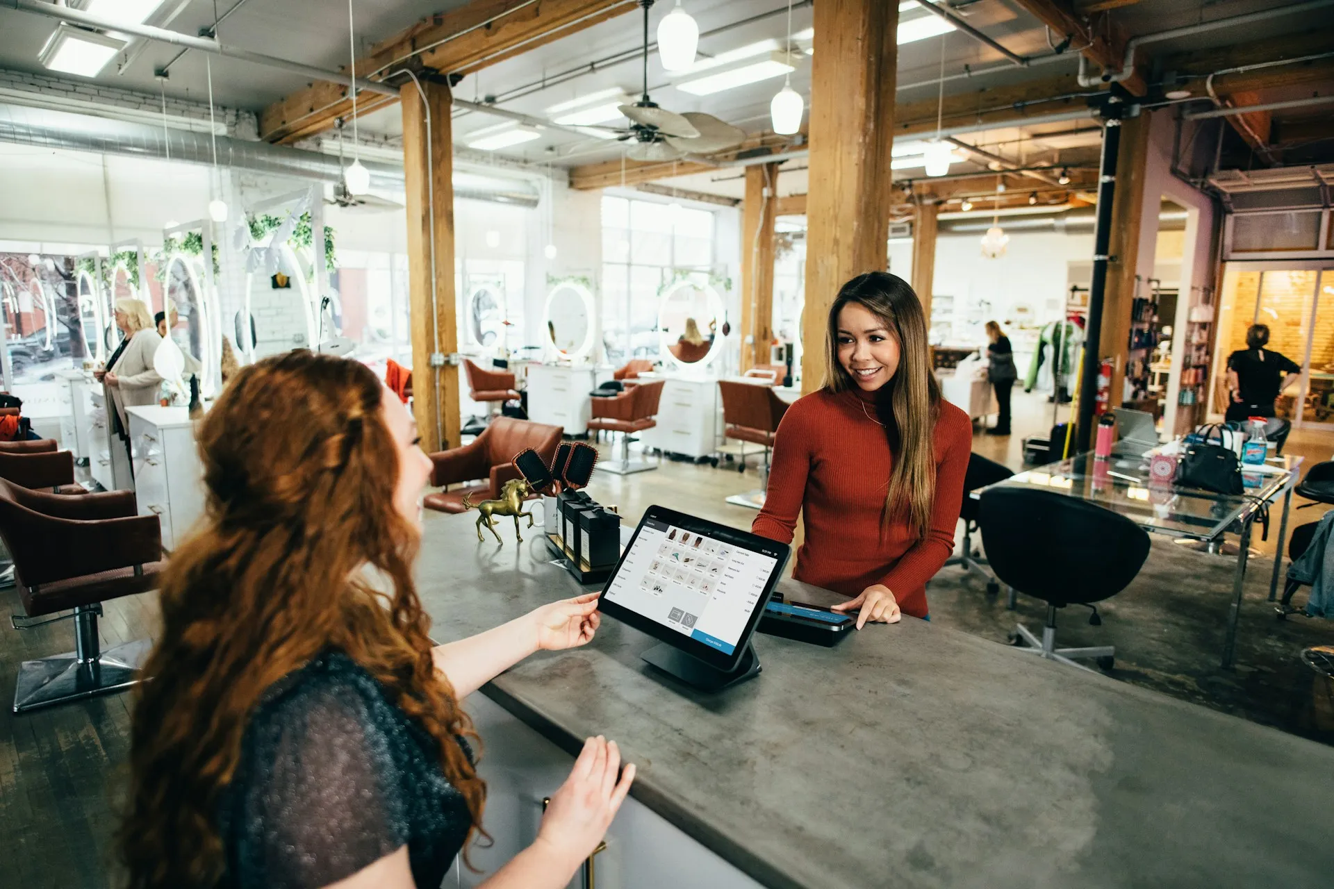 A receptionist in a red sweater smiles at a customer across a reception desk in a modern salon. The receptionist is using a tablet device, likely for scheduling or transactions.