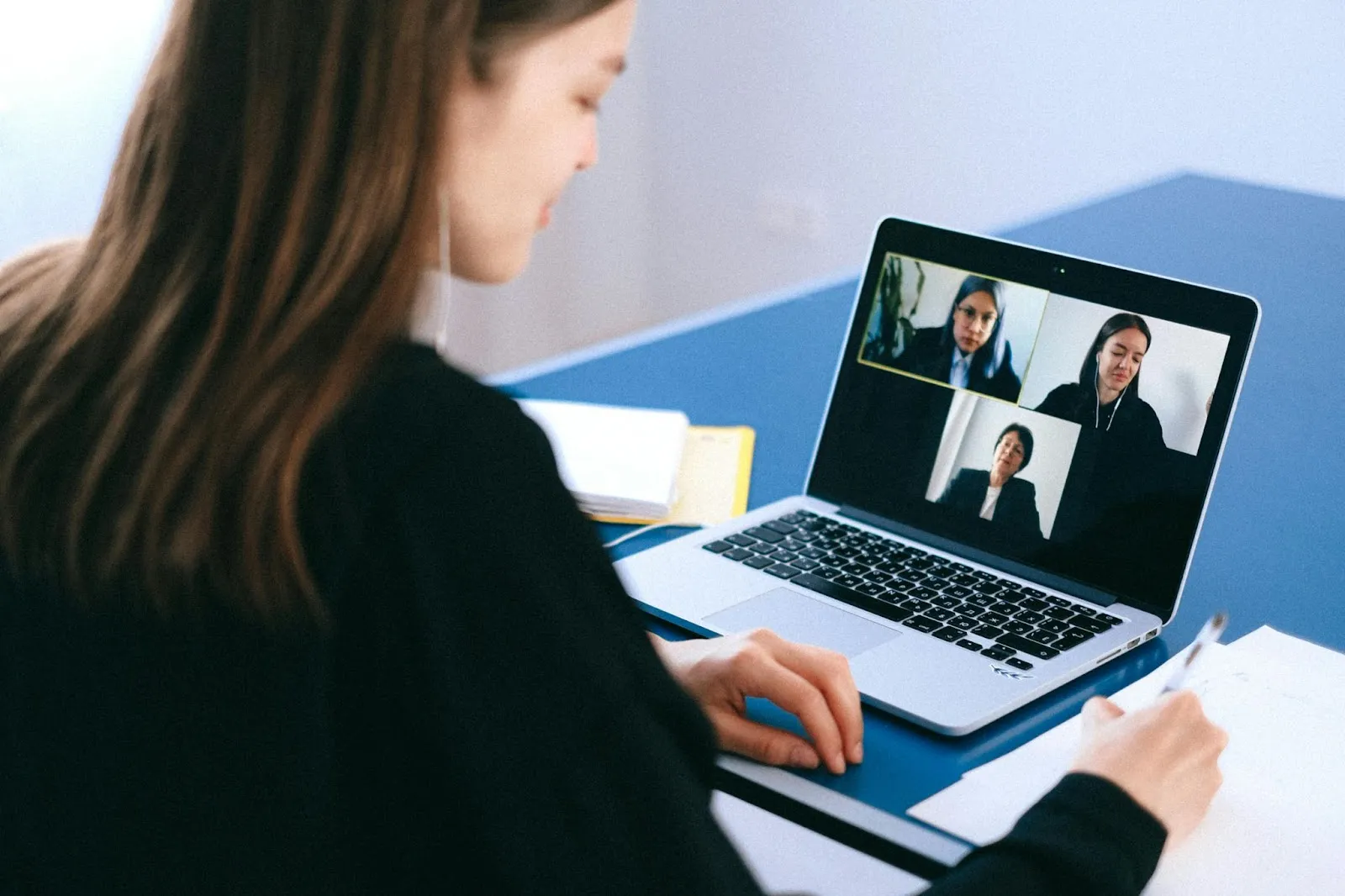 Woman attending a virtual team meeting on her laptop with multiple colleagues visible on a video call, taking notes during the online conference.