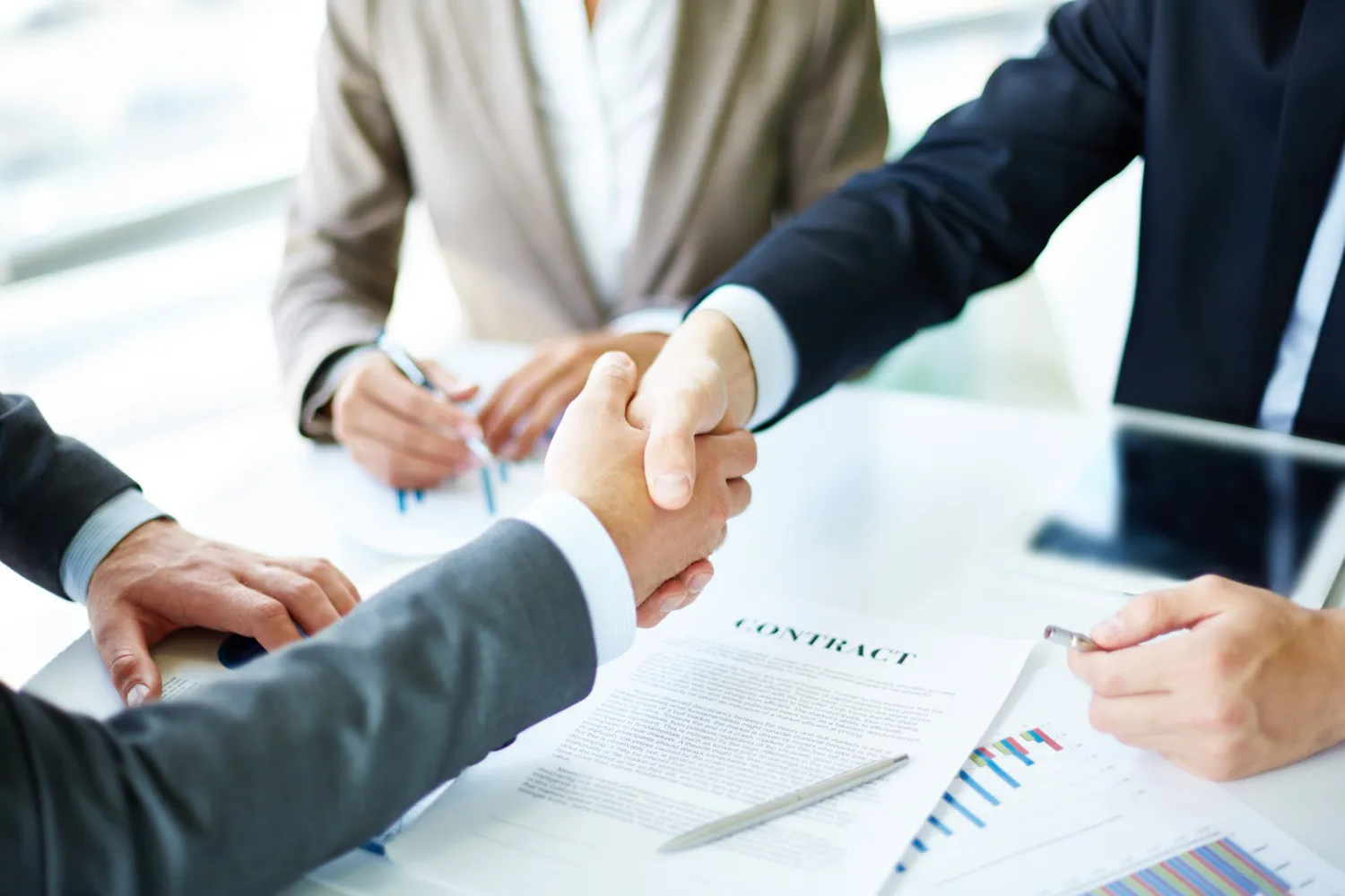 Two business professionals shaking hands over a signed contract at a meeting, with a third person taking notes and reviewing documents in the background.