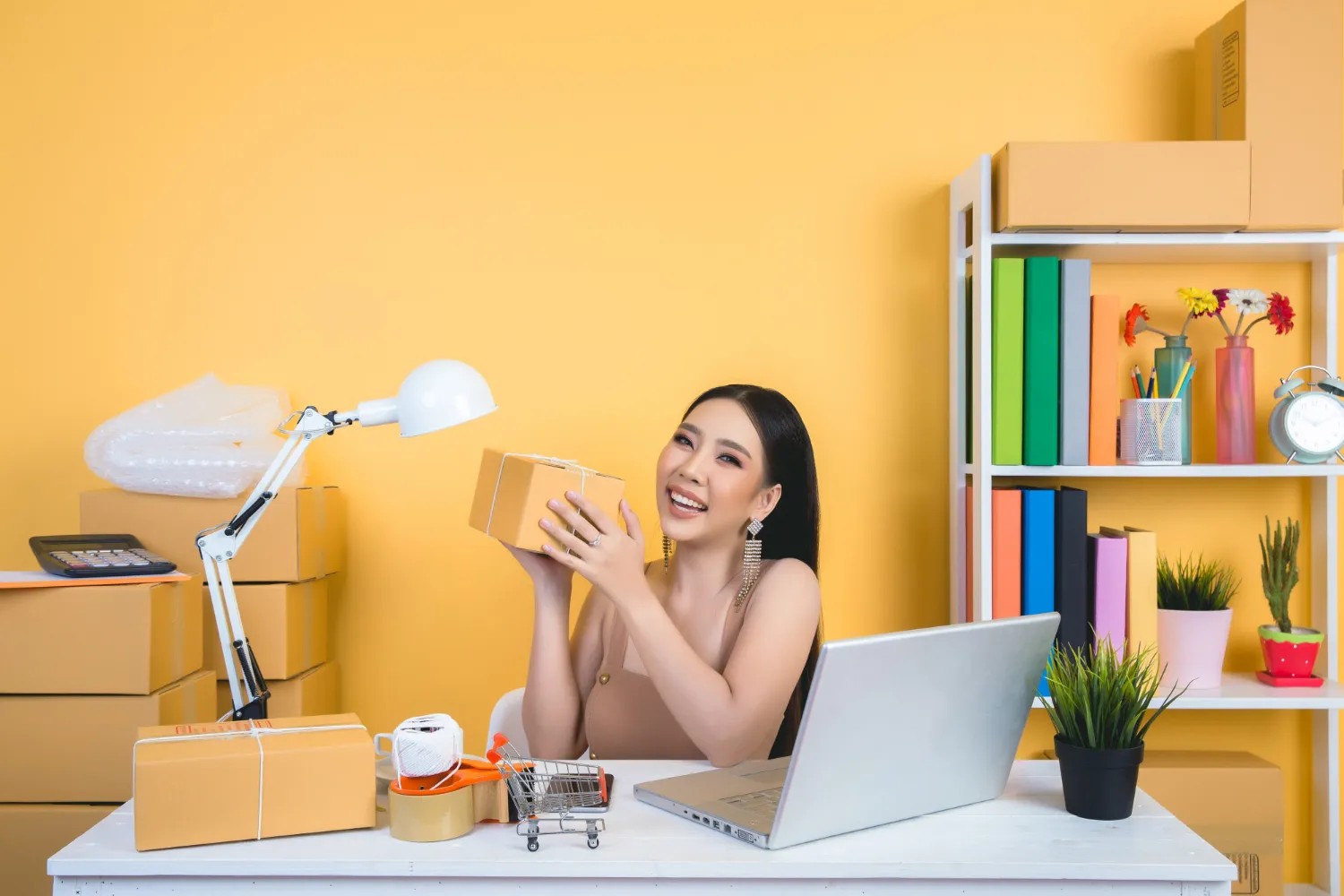 Person working at desk with a laptop, holding a cardboard package, surrounded by packing materials and colorful office supplies.