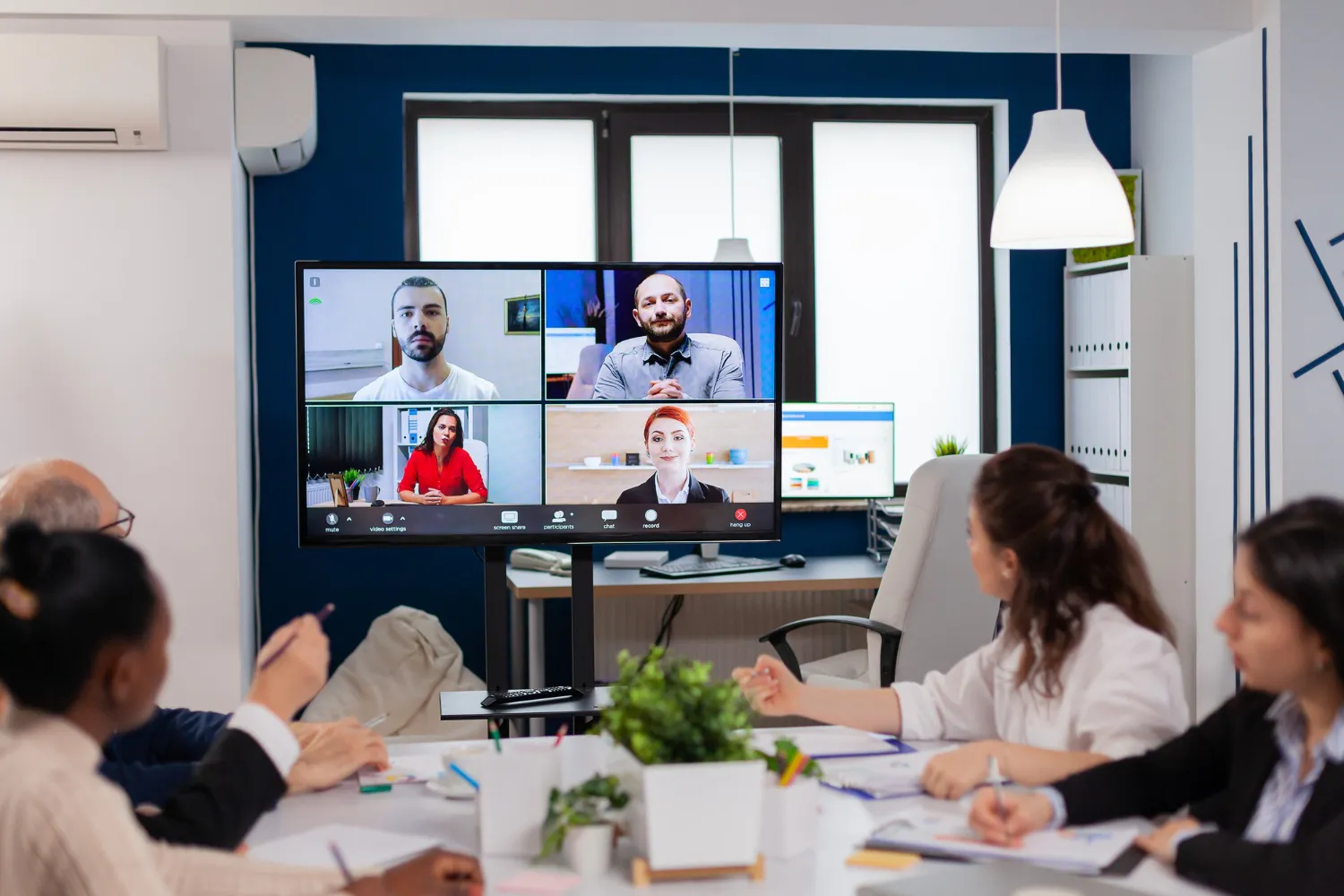 Team of professionals in an office participating in a video conference with four remote colleagues displayed on a large screen.