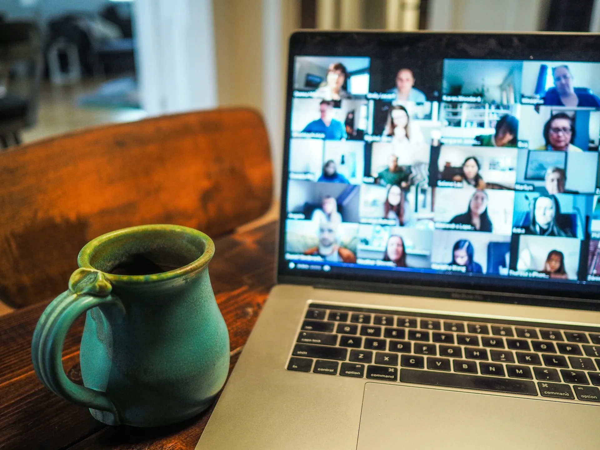 Laptop displaying video conference grid with multiple participants, green ceramic coffee mug on wooden table, remote work setup