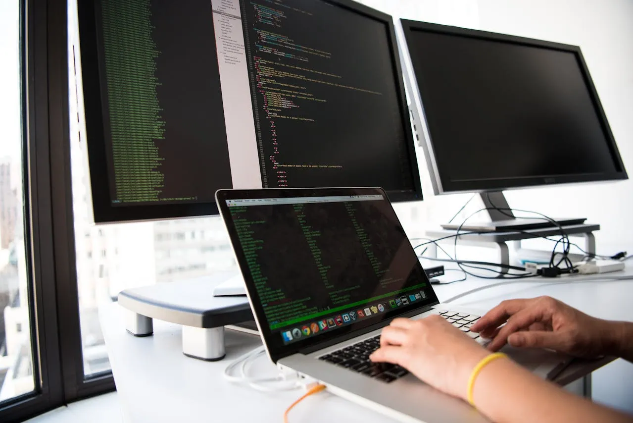 A developer writing code on a laptop with multiple monitors displaying programming code in a modern workspace.