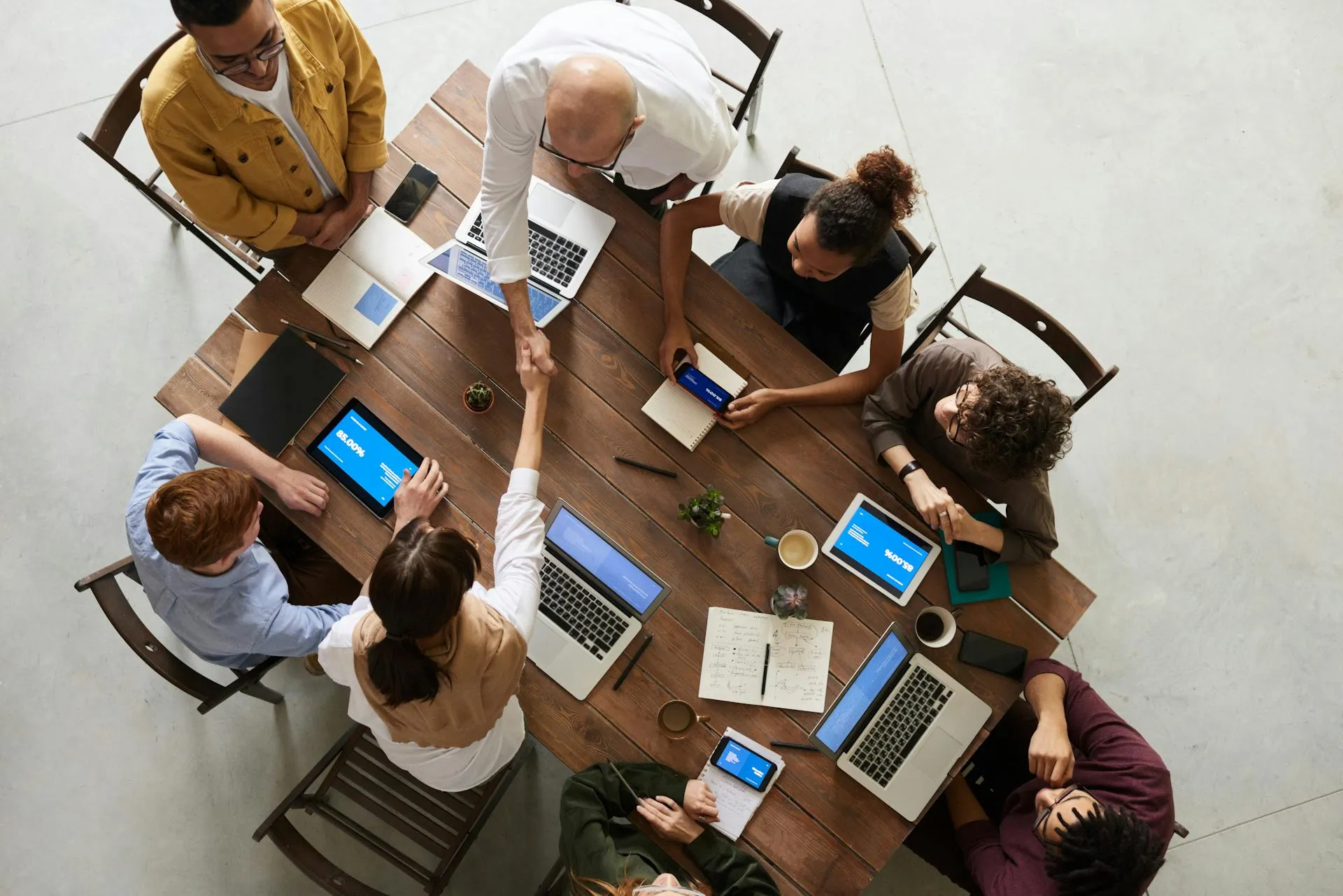 A diverse group of professionals collaborating around a wooden table with laptops and notebooks, shaking hands over a business agreement.