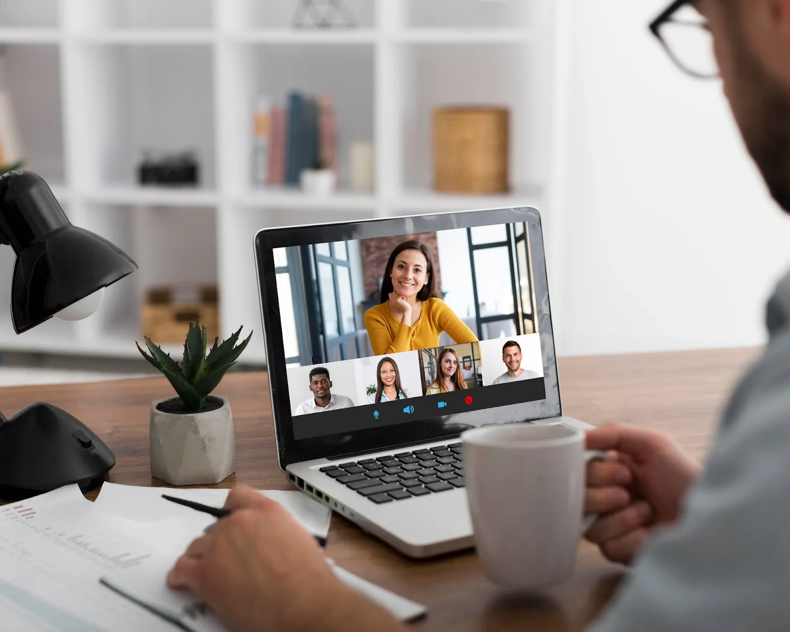 Person participating in video call with multiple team members visible on laptop screen, desk with lamp and plant