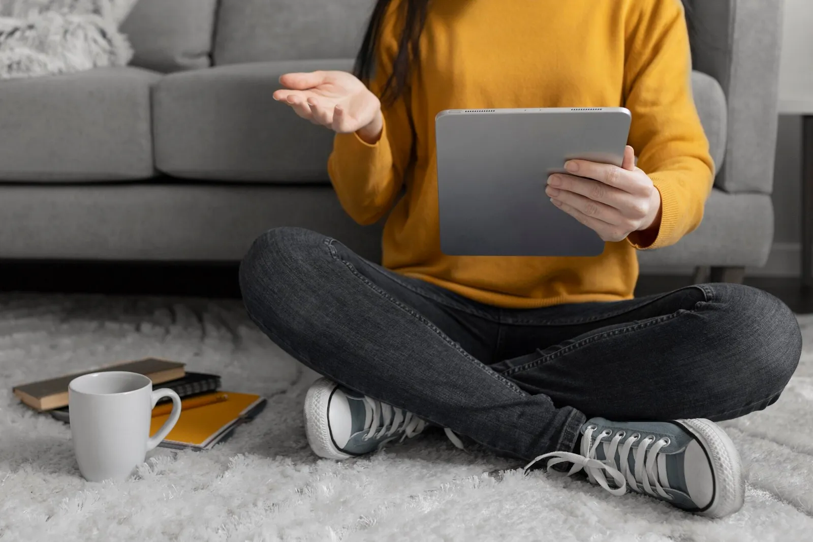 Person in yellow sweater using tablet while sitting on floor, with coffee mug and notebook nearby