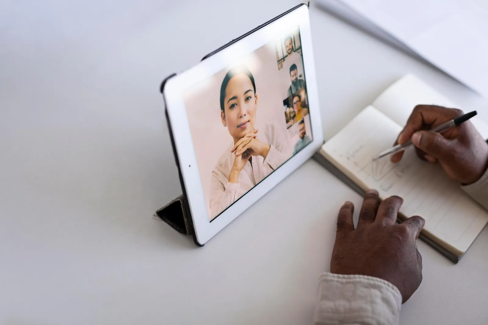 Hands taking notes during video call displayed on tablet, showing woman speaking and other participants