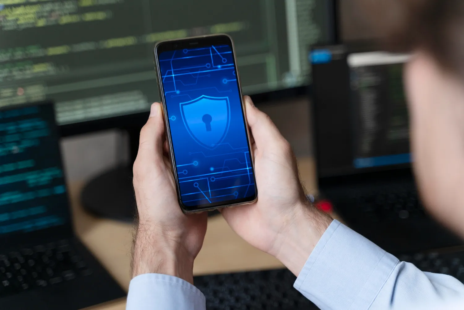 Person holding smartphone displaying blue shield cybersecurity icon, with computer code in background, illustrating mobile device protection and data security.