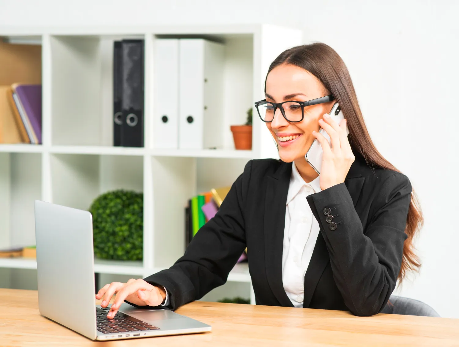 Smiling businesswoman in a black suit using a laptop and talking on a smartphone in a modern office environment.