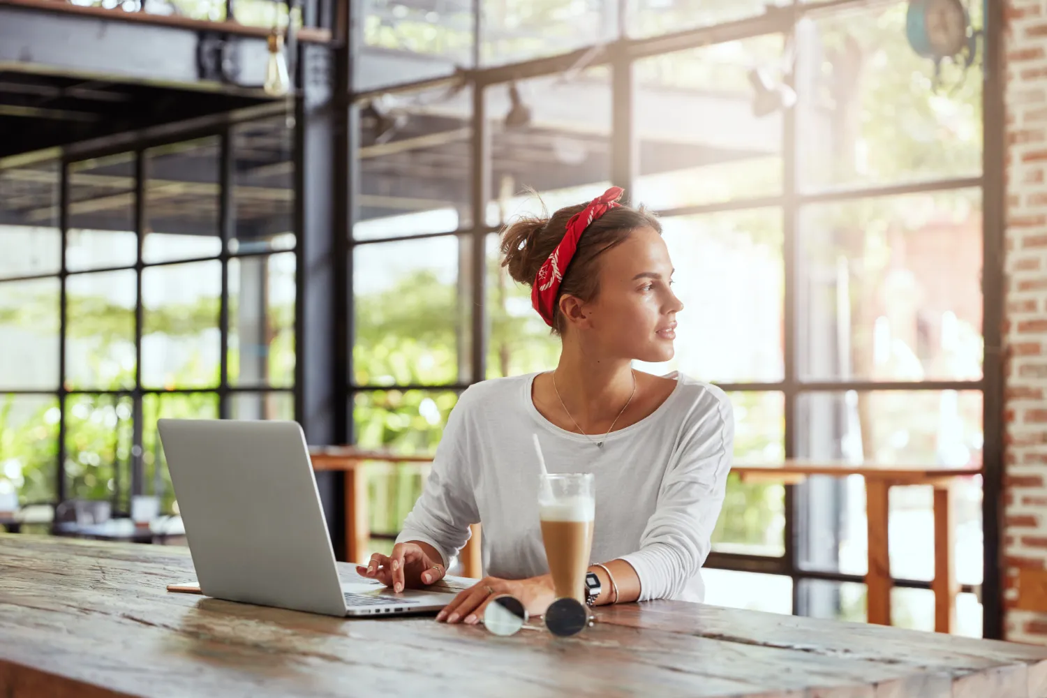 Young woman with red headband working on laptop in sunny cafe, looking thoughtful. Coffee and sunglasses on wooden table. Large windows with greenery visible outside.