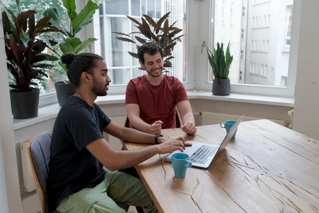 Two professionals discussing work at wooden desk with laptops and plants.